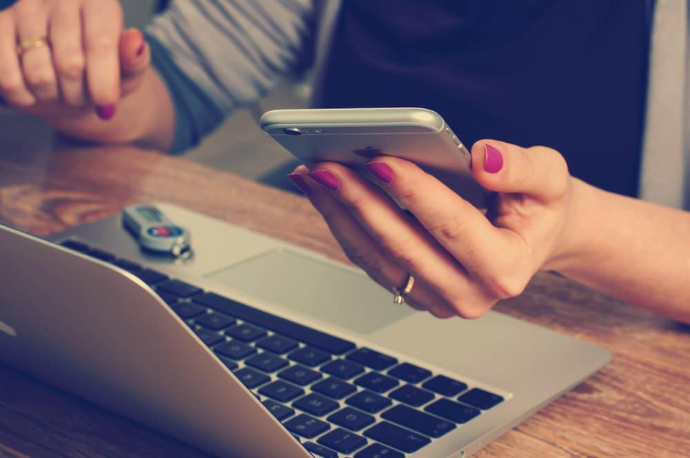 Photo of a person wearing nailpolish looking at their mobile phone with a laptop