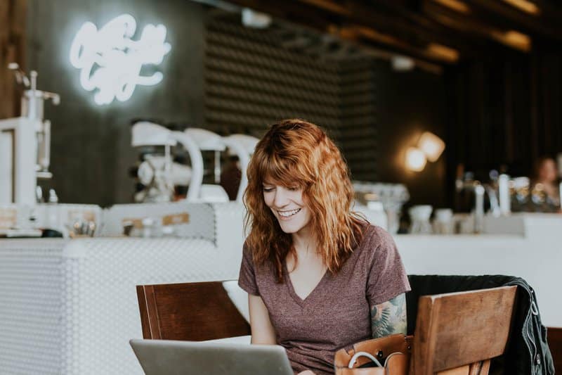 A photo of a red-headed woman sitting in a cafe smiling at a laptop