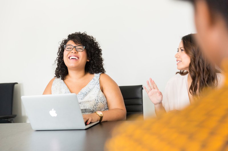 Two women sitting at a conference table, one woman with curly hair and glasses is typing on a laptop adn smiling while the woman on her right speaks to her.