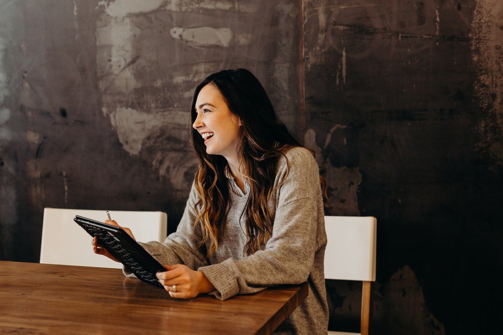 woman with long brown hair sat at a table holding a notepad and pen smiling