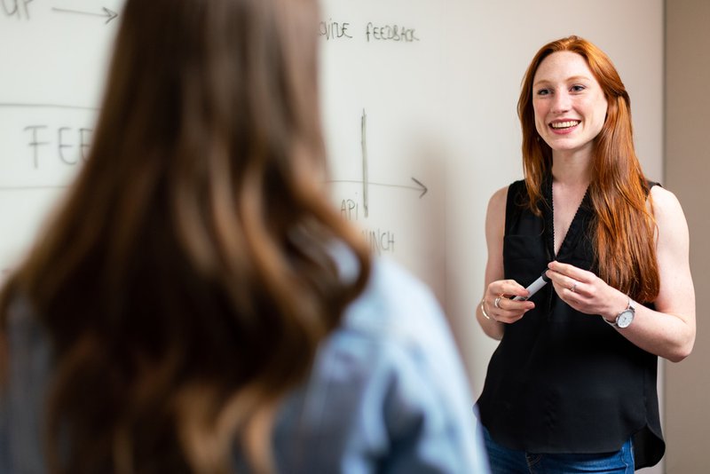 woman in black sleeveless top standing at a whiteboard smiling at a team member