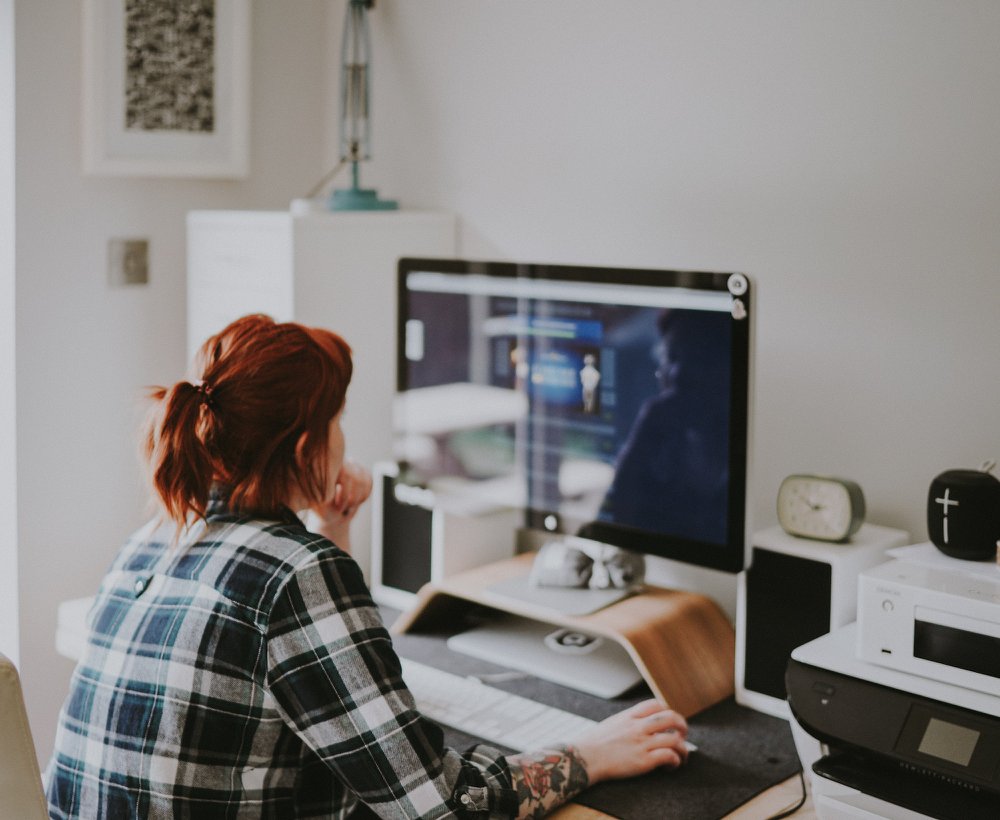 a woman with red hair and a ponytail who works as a Virtual Assistant is sat facing a computer at a desk