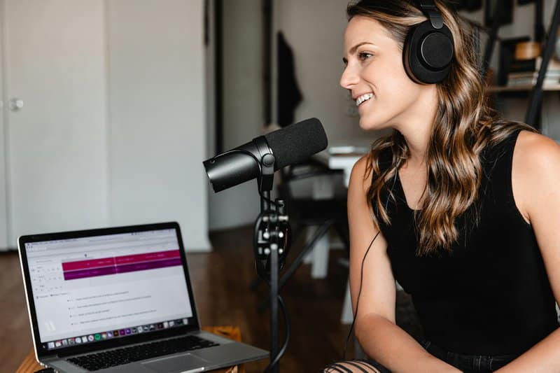 woman wiht blonde hair sitting at a microphone and laptop with headphones on to record a podcast