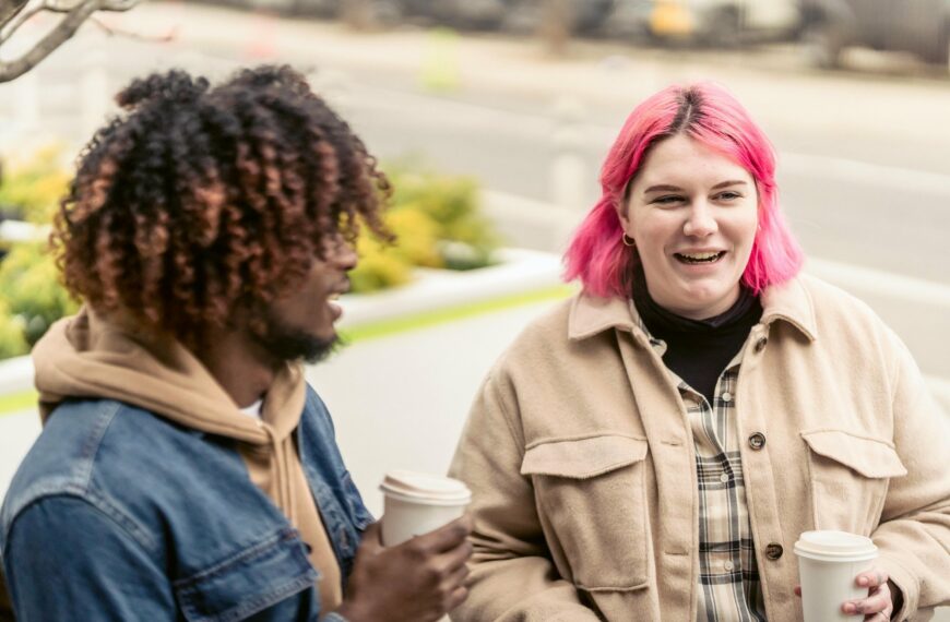 image of a two friends sharing a coffee one is a person with natural hair and the other has bright pink hair, they are both showing, illustrating Virtalent's commitment to being a Mindful Employer