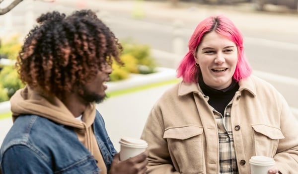 image of a two friends sharing a coffee one is a person with natural hair and the other has bright pink hair, they are both showing, illustrating Virtalent’s commitment to being a Mindful Employer