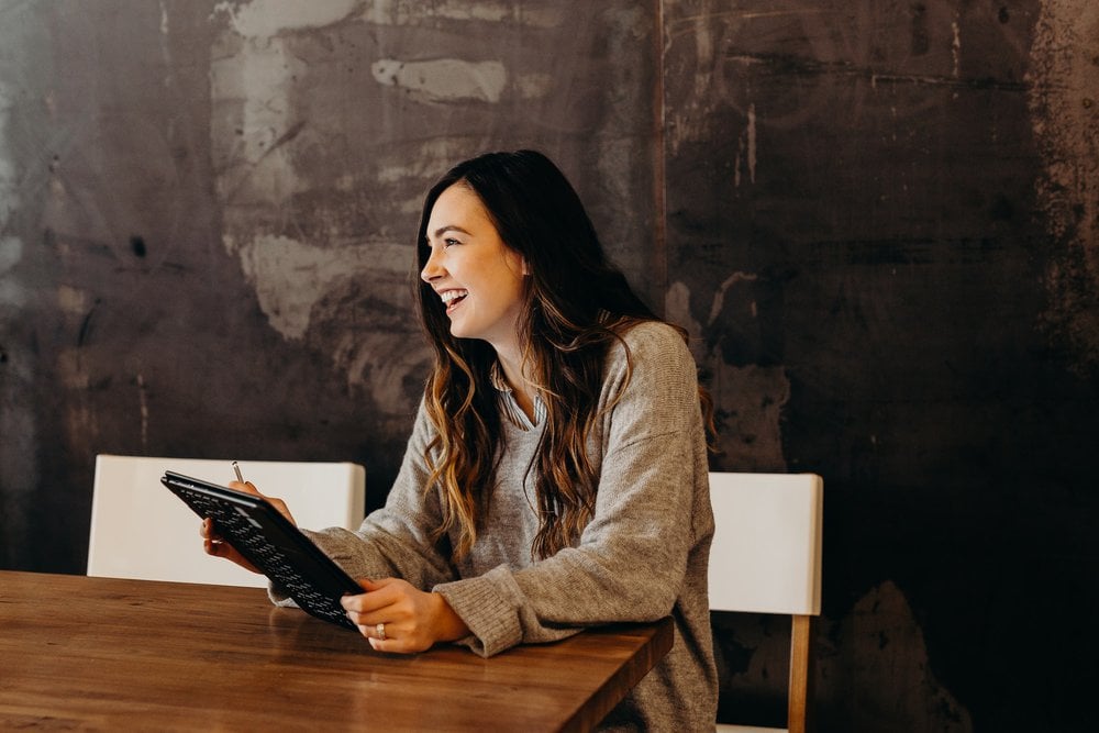 woman with long brown hair sat at a table holding a notepad and pen smiling